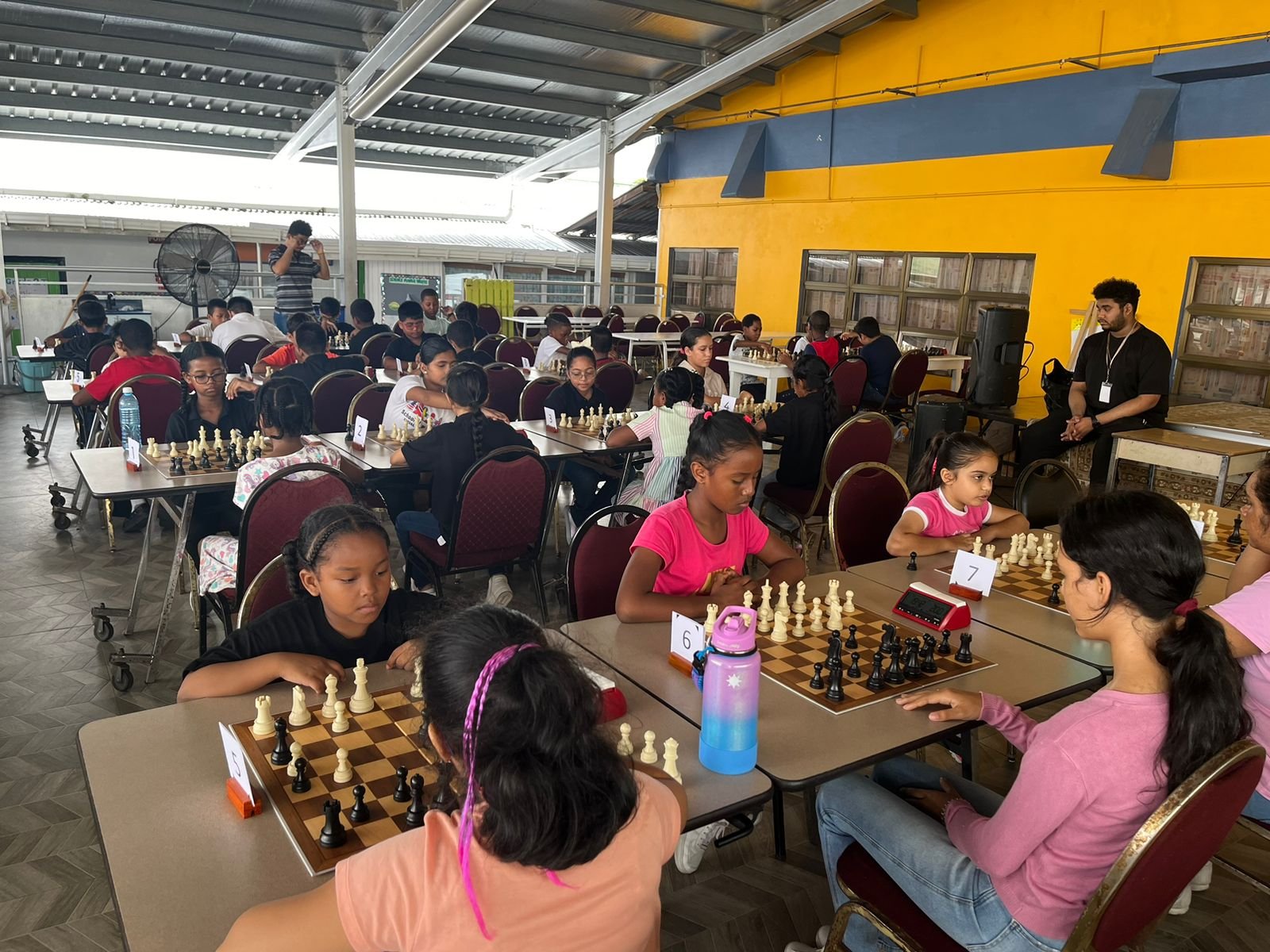 A group of children sit at a wooden desk. They are playing chess.